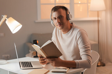 Poster - Young man with headphones, book and laptop studying online at home late in evening