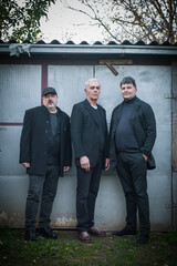 Group of three urban and handsome fashionable artist guys pose in black elegant suits outdoor