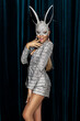 Elegant Woman is wearing Carnival Mask and sequin silver suit and is posing on dark velvet background. Masquerade. Bunny mask. Party costume.