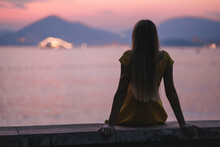 A Young Beautiful Woman In A Dress Sits On The Beach At Sunset And Looks At The Setting Sun, Lonely Girl Dreams Of Love.