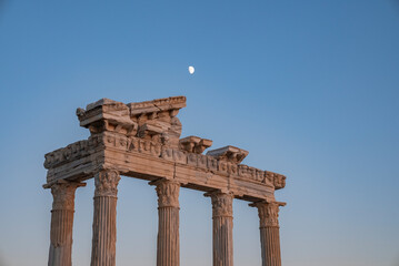 Wall Mural - Low angle view of old ruins of Roman Temple of Apollo with moon in clear blue sky in the background at dusk in Side, Turkey