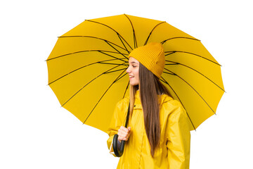 Wall Mural - Teenager girl with rainproof coat and umbrella over isolated chroma key background laughing in lateral position