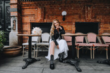 A Beautiful Curly Smiling Blonde Bride, Biker, Rock Lover In A Black Leather Jacket, White Dress With A Bouquet Of Reeds Sits On Chairs In The City On The Street. Wedding Photography, Portrait.