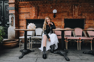 A beautiful curly smiling blonde bride, biker, rock lover in a black leather jacket, white dress with a bouquet of reeds sits on chairs in the city on the street. Wedding photography, portrait.