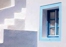 A Famous Blue Painted Window Of The Greek Islands- Santorini. A Vibrant Frame Looks At The White-washed Walls And Stairs Of The Traditional Cave Homes. The Summer Sun Creates Tranquil Artistic Shadows