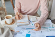 Cropped shot of unknown woman poses at home office makes calculations of utility payments writes down notes in spiral notebook surrounded by paper bills and cup of coffee calculates domestic expenses