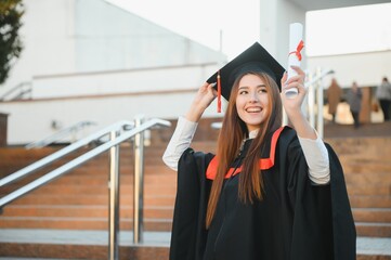 Happy cute brunette caucasian grad girl is smiling. She is in a black mortar board, with red tassel, in gown, with nice brown curly hair, diploma in hand.