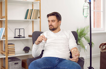 Tired Young Man Sitting In Armchair At Home, Receiving Medication Infusion Through Intravenous Drip, Holding Glass, Taking Medicine, Drinking Water And Thoughtfully Looking Away