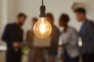 Glowing light bulb and business people over blurred background. Hanging light bulb on foreground, team of business people discussing ideas and brainstorming. Bright idea and creativity concept