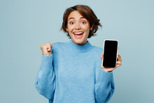 Young Smiling Happy Woman In Knitted Sweater Hold In Hand Use Mobile Cell Phone With Blank Screen Workspace Area Do Winner Gesture Isolated On Plain Pastel Light Blue Cyan Background Studio Portrait