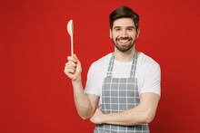 Young Smiling Cheerful Happy Fun Male Housewife Housekeeper Chef Cook Baker Man Wearing Grey Apron Hold In Hand Wooden Spoon Look Camera Isolated On Plain Red Background Studio. Cooking Food Concept.