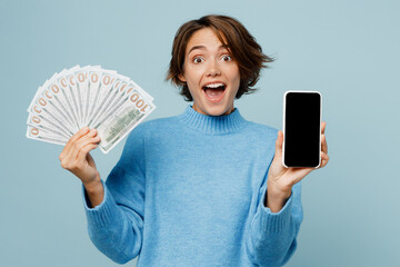 Wall Mural - Young surprised fun woman wear knitted sweater use mobile cell phone blank screen workspace area hold fan of cash money in dollar banknotes isolated on plain pastel light blue cyan background studio.