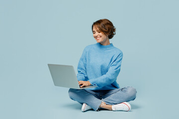 Full body young smiling happy fun cool IT woman wear knitted sweater hold use work on laptop pc computer isolated on plain pastel light blue cyan background studio portrait. People lifestyle concept.