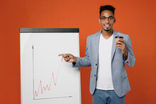 Young Fun Employee Business Man Corporate Lawyer In Formal Grey Suit Shirt Glasses Work In Office Use Flipchart Show Graph On Marker Board Drink Coffee Isolated On Plain Red Orange Background Studio