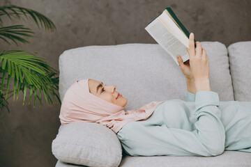 Wall Mural - Young calm muslim woman wearing hijab casual clothes reading book lying on sofa couch stay at home flat rest relax spend free spare time in living room indoor. People middle eastern uae islam concept.