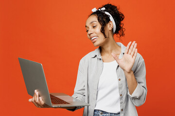 Smiling young IT woman of African American ethnicity she wears grey shirt headband hold use work on laptop pc computer waving hand isolated on plain orange background studio People lifestyle concept.