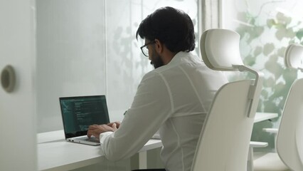 Over the shoulder view of focused qualified IT developer working in bright office writing code on laptop. Attractive professional Indian man programming using technologies working in company.