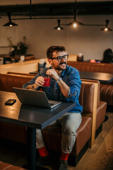 Wall Mural - Bearded smiling Caucasian freelancer sitting in a cafe, drinking a fresh smoothie and using a laptop.