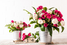 Many White And Red Roses In Two Metal Decorative Buckets On A Marble Table Against A White Brick Wall. Wooden Calendar With A Valentine's Day Holiday.