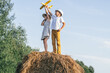 Little girl and boy together launch yellow toy airplane standing at top of haystack. Children on hayrick from below view