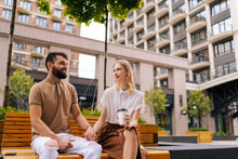 Low-angle View Of Happy Loving Man And Woman Enjoying Coffee, Smiling Talking Sitting On Bench In City Street. Portrait Of Loving Couple Relaxing Outdoors Drinking Coffee During Romantic Date.