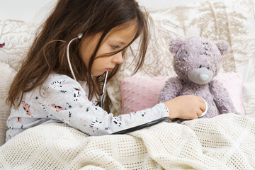 Serious little girl in sleepwear sitting under blanket and listening to lung of teddy bear with stethoscope. Portrait of child, side view. Kid and toy have bed rest. Home patient treatment.