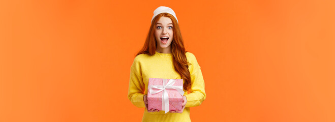 Wall Mural - Surprised and excited happy girl unwrapping gifts on christmas eve, new year celebration, smiling amused and thrilled, holding wrapped box present, wearing winter beanie, orange background