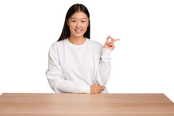 Wall Mural - Young asian woman sitting on a table isolated smiling cheerfully pointing with forefinger away.