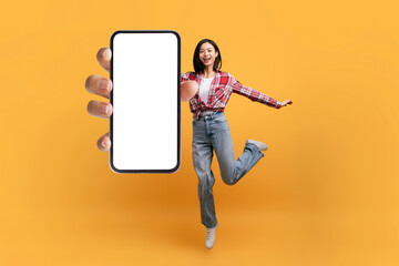 Full length of happy asian woman jumping and showing cellphone with empty space for mobile app or website, mockup