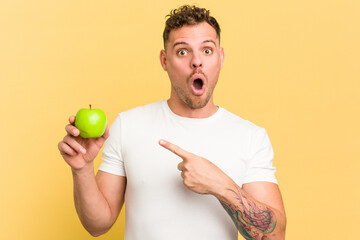 Young caucasian man eating an apple isolated pointing to the side