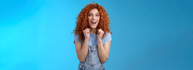 Cheerful optimistic lucky redhead curly-haired attractive woman cheering clench fists joyfully smiling broadly cheering watching game supportive encourage keep up motivated blue background