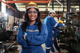 Fototapeta  - Portrait of professional Black female worker in protective safety uniform and helmet looking at camera, arms crossed and smiling, with engineers team behind her in a metalwork manufacturing factory.