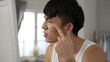 © PRPicturesProduction - closeup view of an upset asian young man discovering pimple on face and trying to get rid of it in front of the mirror in the morning at his bed chamber.