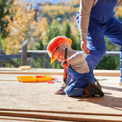 Wall Mural - Father with toddler son building wooden frame house. Male builders hammering nail into plank on construction site, wearing helmet and blue overalls on sunny day. Carpentry and family concept.