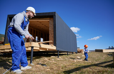 Wall Mural - Father with toddler son building wooden frame house. Male builder and kid playing with tape measure on construction site, wearing helmet and blue overalls on sunny day. Carpentry and family concept.