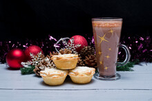 Tall Glass Of Hot Chocolate SittingTall Glass Of Hot Chocolate Sitting Beside The Christmas Decorations Ready To Be Hung On The Tree With Three Freshly Made Christmas Mince Pies 