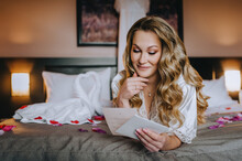 A Beautiful Smiling Curly Blonde Bride In Lingerie, A Dressing Gown Lies In The Morning In A Room On A Bed In Rose Petals, A Bedroom, Reading An Oath From A Paper Envelope. Wedding Photography.