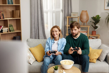 Young Couple Playing Video Games. Boyfriend And Girlfriend Sitting On Couch In Living Room Enjoying Playing Video Games And Spending Time Together. Enjoying Moment On Weekend Concept 