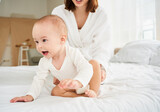 Fototapeta  - A laughing toddler in a white bodysuit crawls away from mom on the bed.