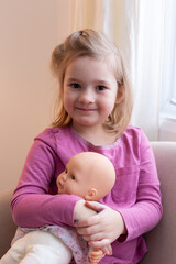 Wall Mural - Vertical view of beautiful smiling little girl with blond hair and brown eyes in pink shirt sitting next to a window holding her doll