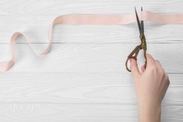 Wall Mural - Woman cutting beige ribbon with scissors at white wooden table, closeup. Space for text