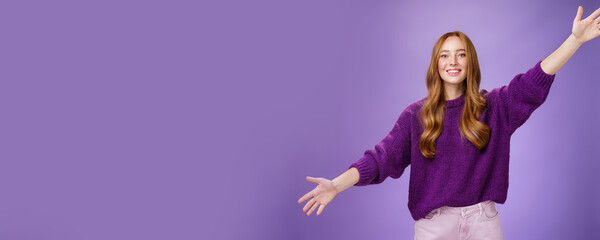 Wall Mural - Girl stretched hands sideways to welcome and greet friend giving warm hug smiling broadly at camera standing joyful wanting cuddle over purple background, wearing violet sweater and pants