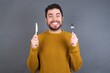 © Jihan - Young handsome caucasian man wearing yellow knitted sweater over gray background holding a fork and knife feeling excited looking front