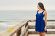© Retamosa - a woman looking at the horizon in a blue dress touching the texture of wooden railing