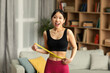 Portrait of positive asian lady in sportswear measuring waist with tape, feeling happy about losing weight at home