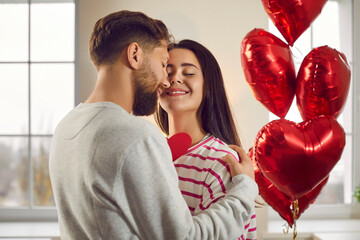 Wall Mural - Happy man and woman celebrate Valentine's Day and make romantic gifts to each other. Indoor shot of a young couple in love hugging, with red heart shaped balloons in the background