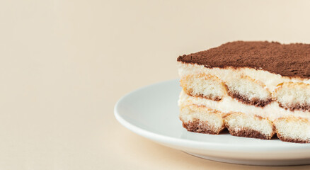 Wall Mural - Homemade tiramisu cake on white plate against beige background. Traditional Italian no-bake dessert made of savoiardi, filled with mascarpone cheese, coffee espresso and sprinkled with cocoa powder