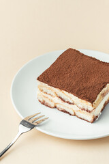Wall Mural - Homemade tiramisu cake on white plate against beige background. Traditional Italian no-bake dessert made of savoiardi, filled with mascarpone cheese, coffee espresso and sprinkled with cocoa powder