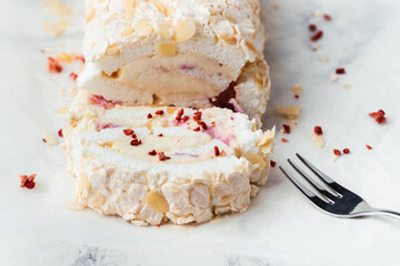 Wall Mural - Cut of homemade meringue roll decorated with almond flakes and freeze-dried red berries on the white marble background. Meringue roulade with whipped cream on a parchment paper