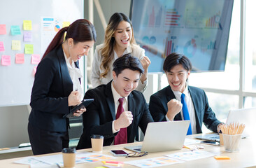 Wall Mural - Millennial Asian young professional successful  businessman in formal suit laughing holding fists up celebrating with female and male businessman colleagues after winning job acheivement deal done